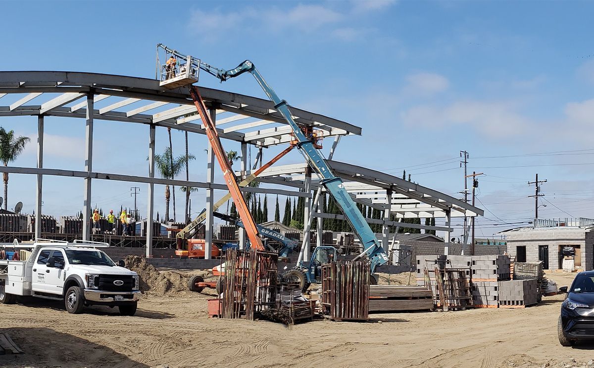 Construction site with steel frame building, two cranes, and parked vehicles on dusty ground
