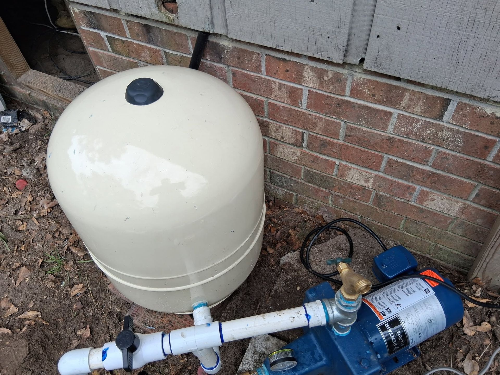 White pressure tank and blue water pump connected by pipes beside a brick wall outdoors