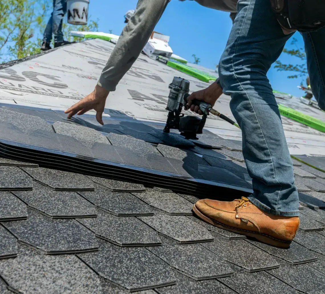 A person in jeans and work boots uses a nail gun to install dark asphalt shingles on a sloped roof.
