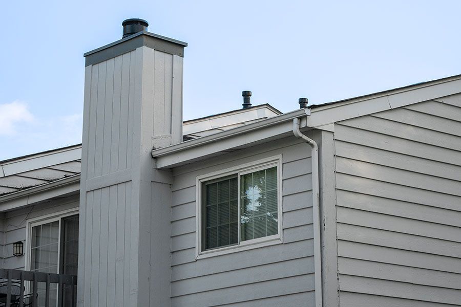 A light gray house exterior featuring a tall chimney stack, horizontal siding, a sliding window, and a roofline against sky.