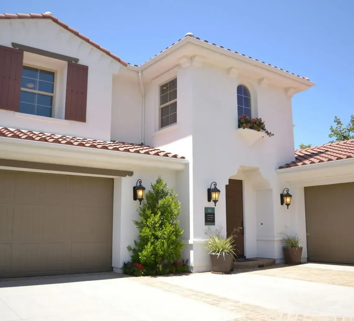 A two-story, white stucco house with a tiled roof, two garage doors, and a front entry under a clear blue sky.