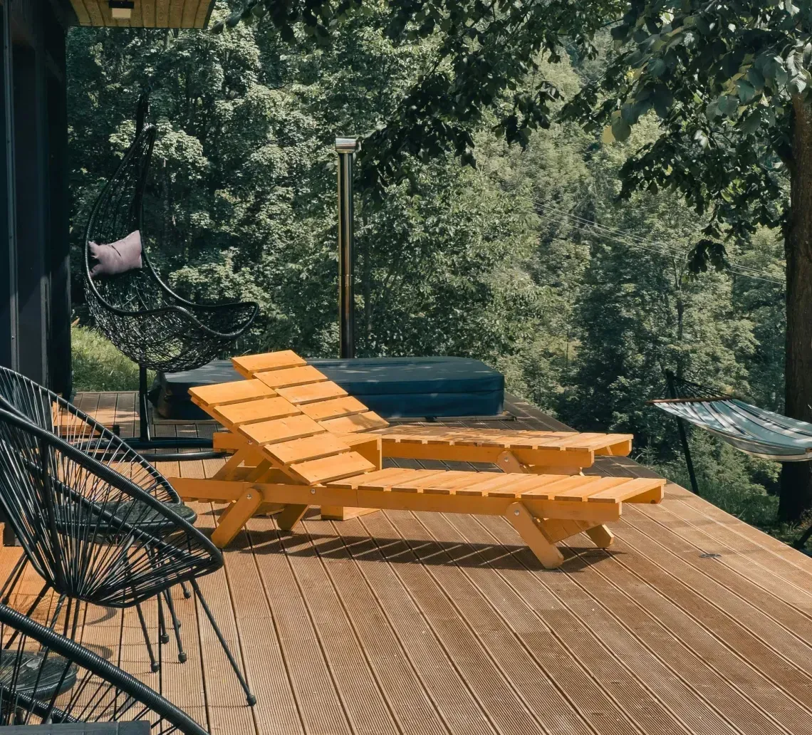 Wooden deck featuring two orange lounge chairs, black metal chairs, and a small hot tub overlooking a lush forest.