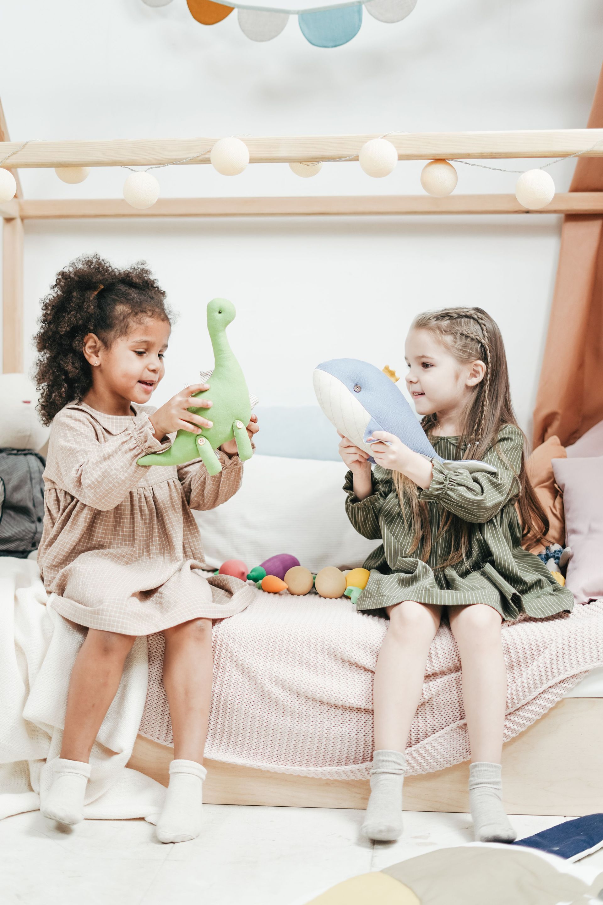 Two children sitting on a bed, playing with stuffed animal toys in a cozy bedroom.