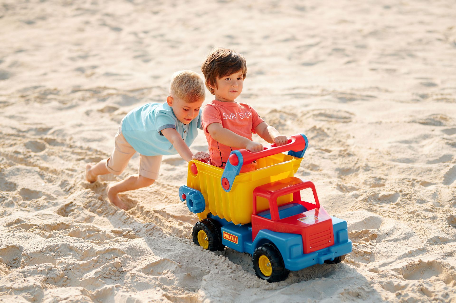 Two children pushing a colorful toy dump truck in the sand at the beach