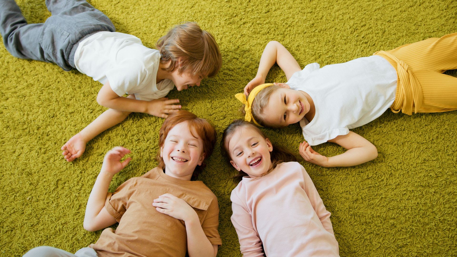 Four children lying on green grass, smiling and relaxing in a circle.