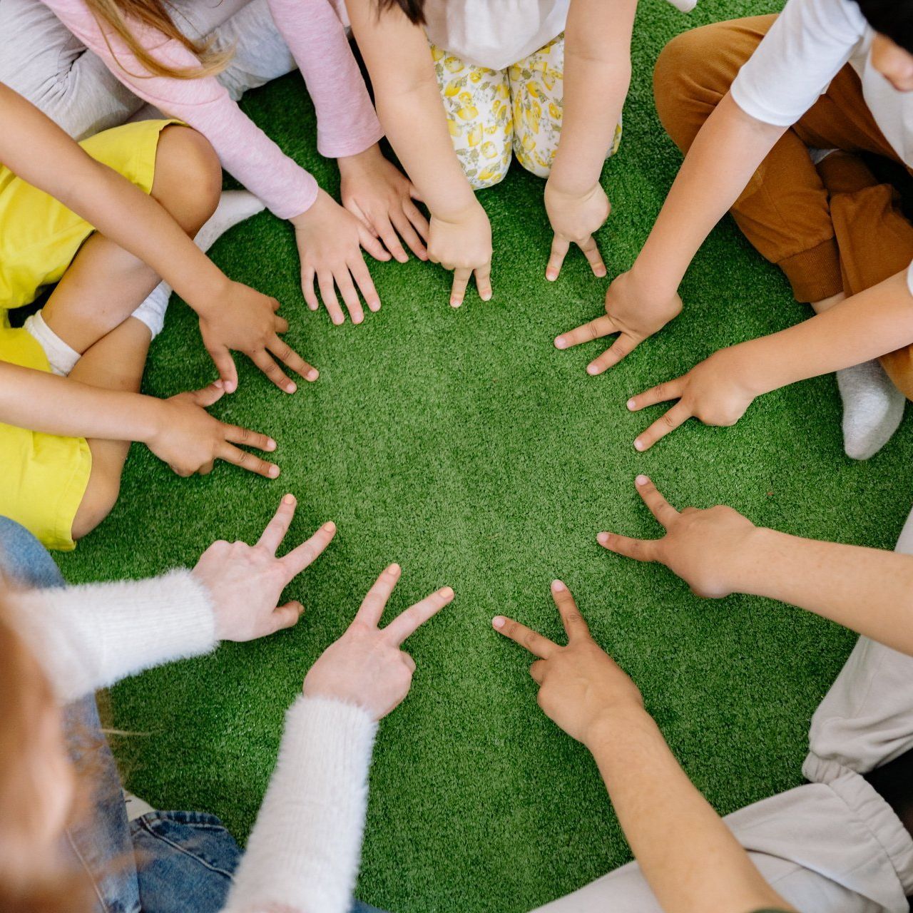 Children’s hands arranged in a circle over green grass, making peace signs and pointing inward