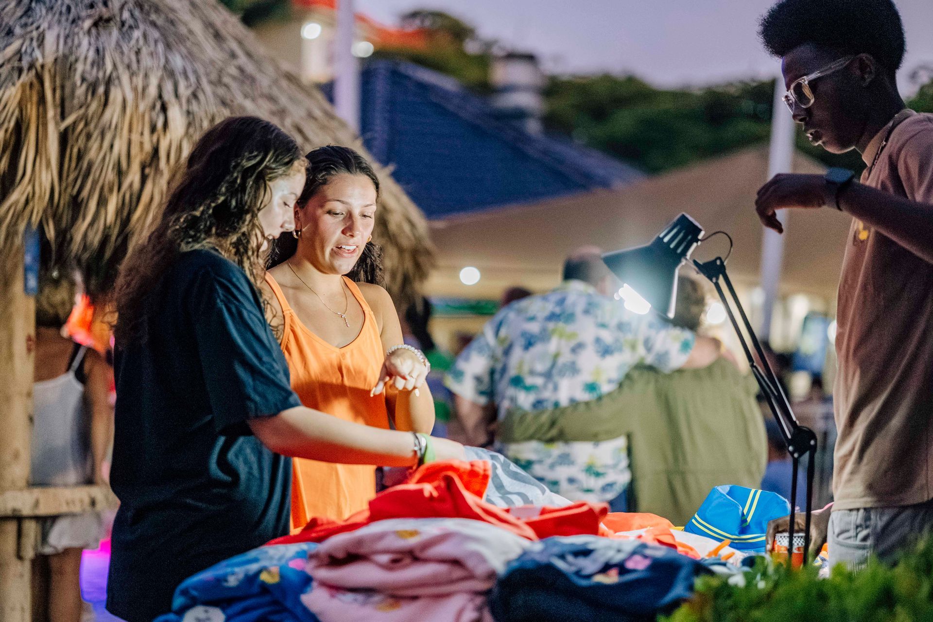 Two women looking at clothing at a stand with a man; outdoor market at dusk.