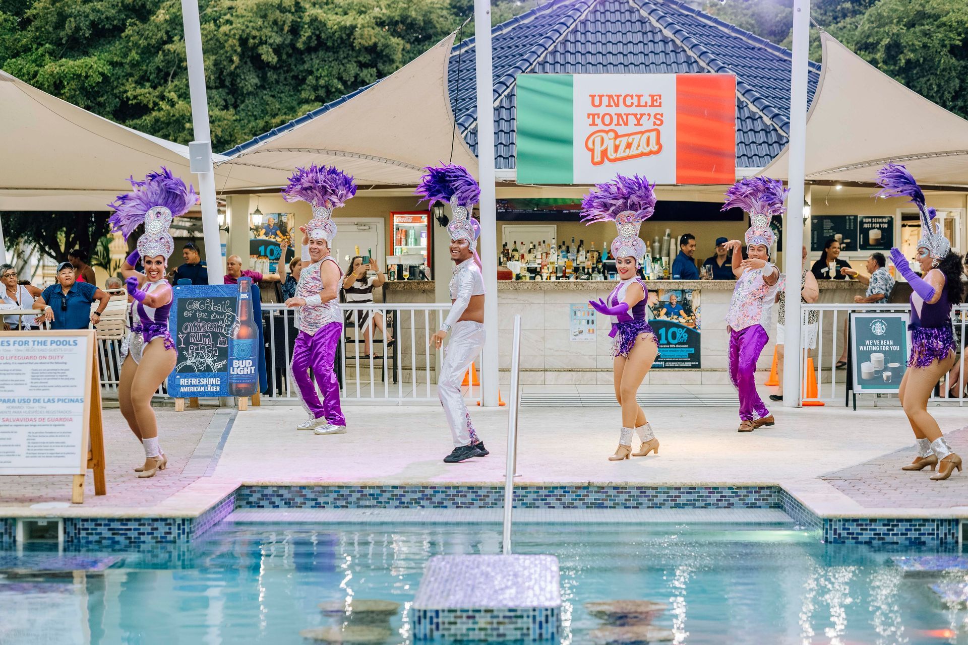 Dancers in purple and gold costumes perform near a pool, with an Italian restaurant sign in the background.