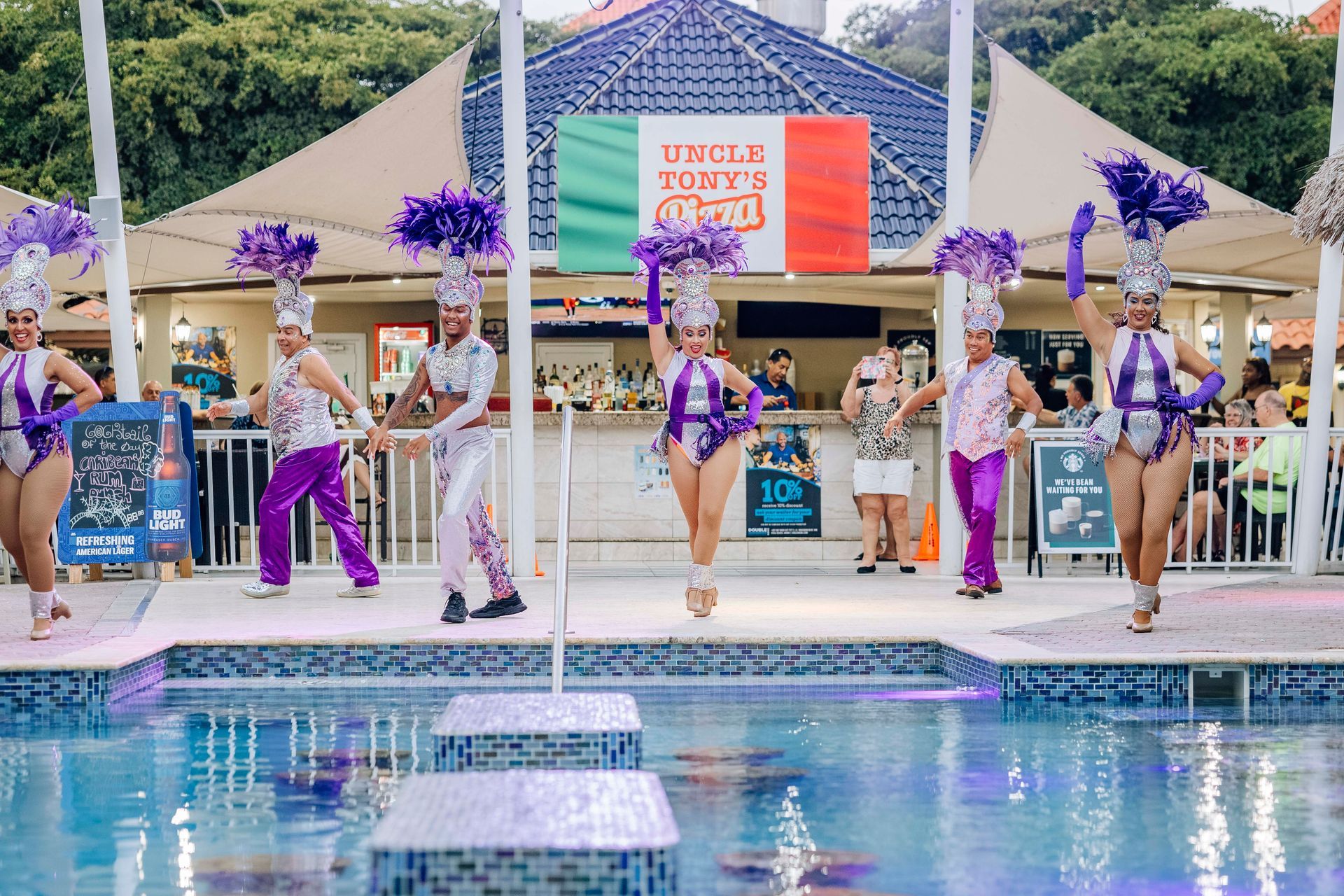 Performers in purple and silver costumes dance by a pool near a bar, 
