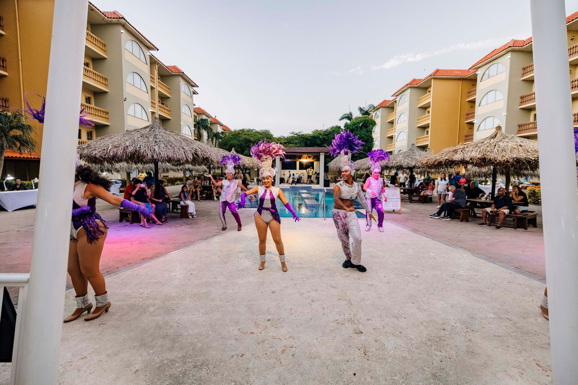 Dancers performing in an outdoor venue; buildings and thatched umbrellas in the background.