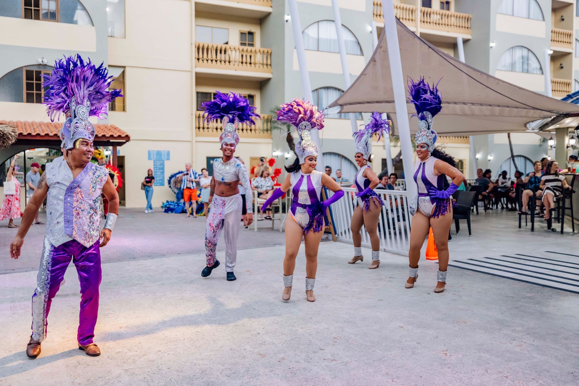 Dancers in vibrant purple costumes with feather headdresses performing outdoors in front of an audience.