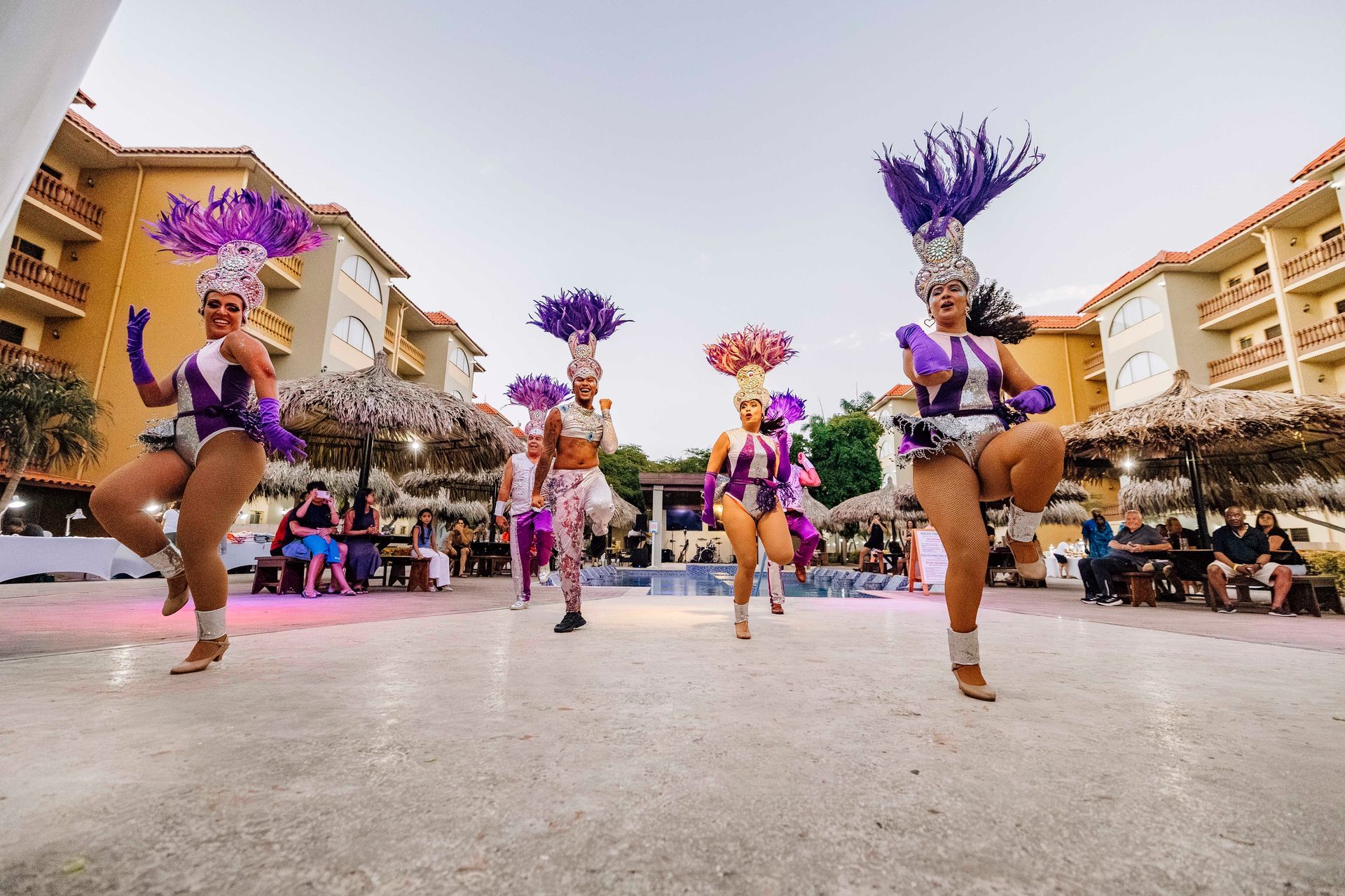 Dancers in purple costumes with feathered headdresses perform outdoors near a resort.