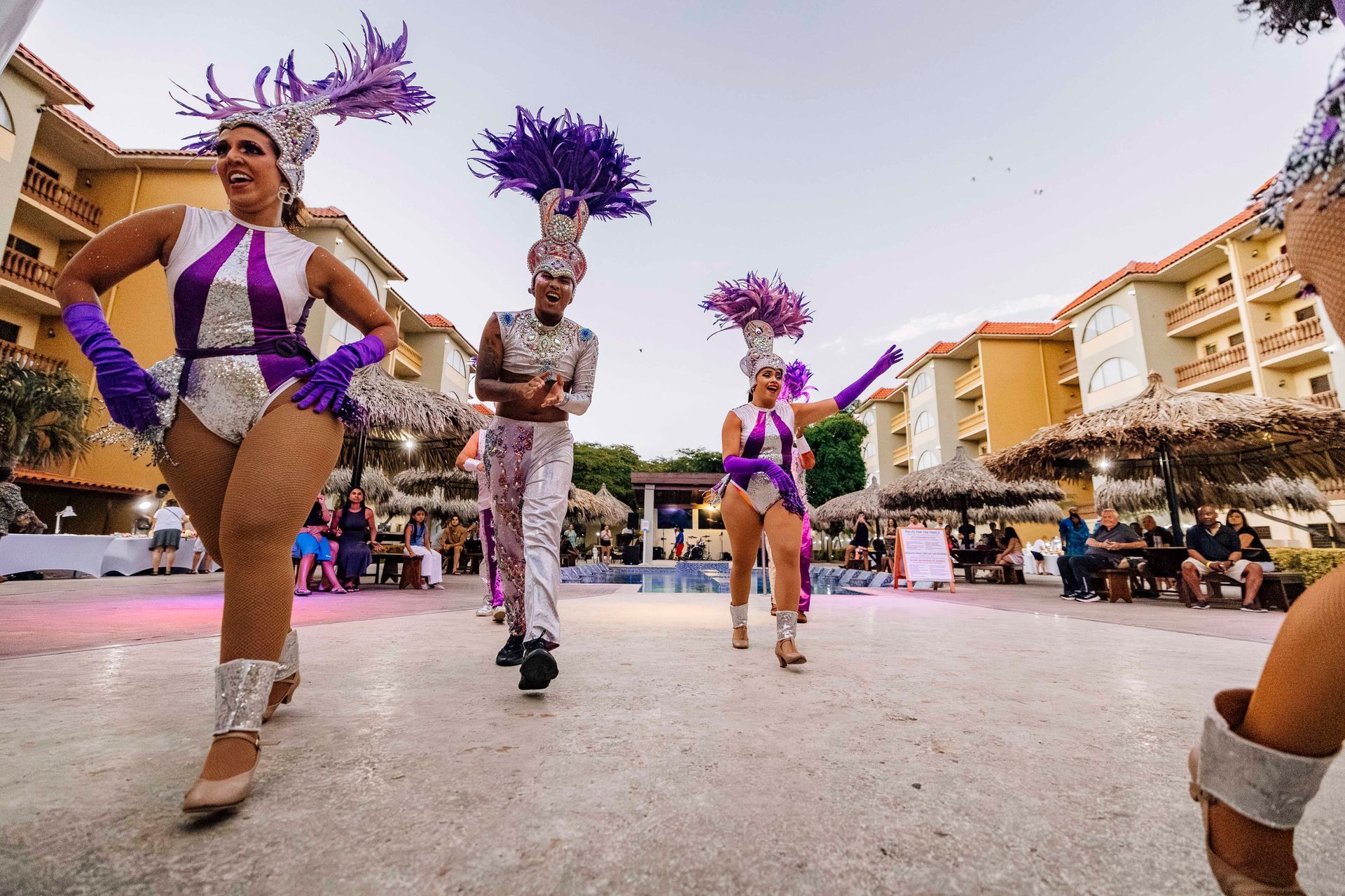 Dancers in purple and white costumes perform outdoors at a resort, smiling, with feather headdresses.
