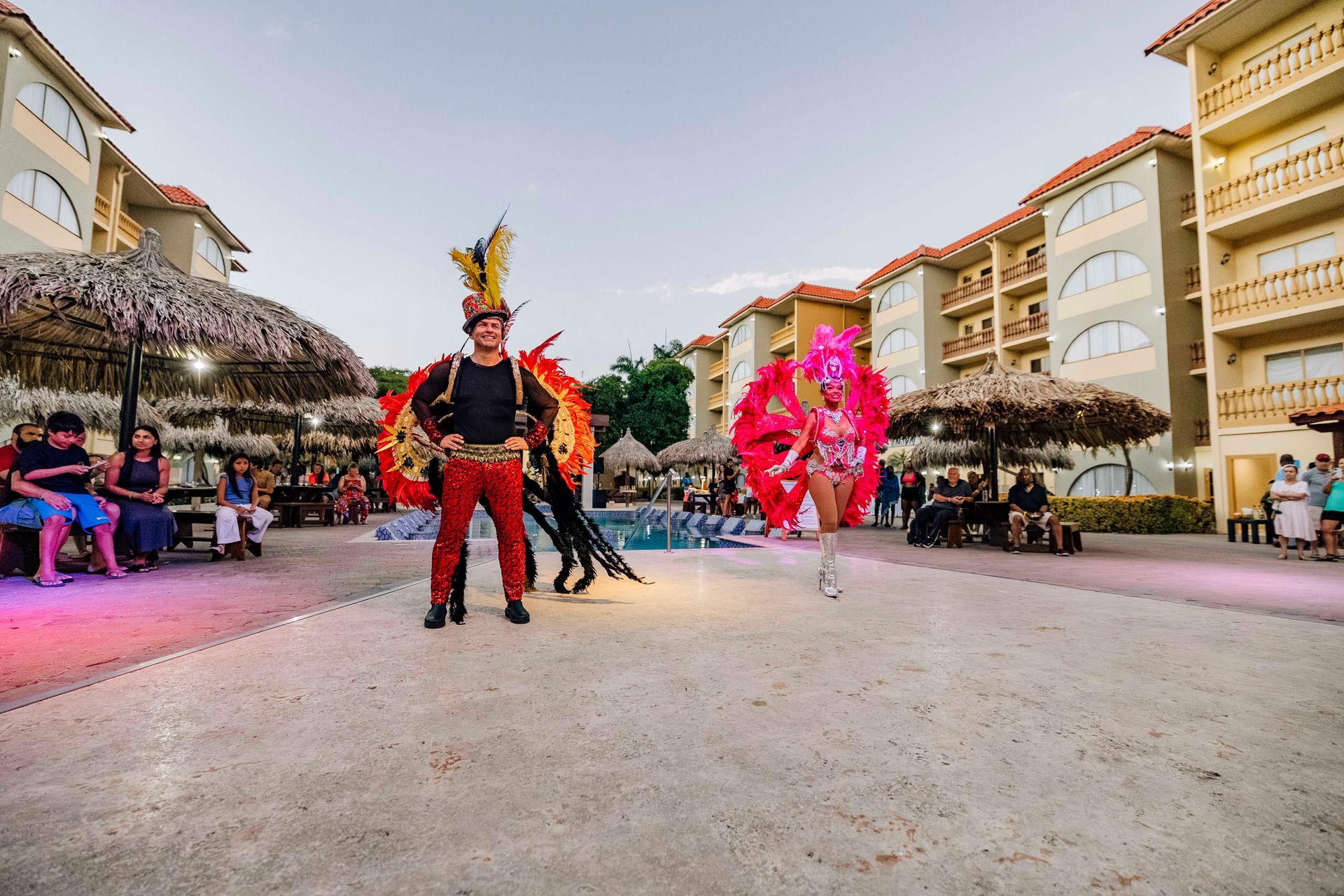 Two people in elaborate costumes perform at an outdoor resort. Buildings and audience visible.