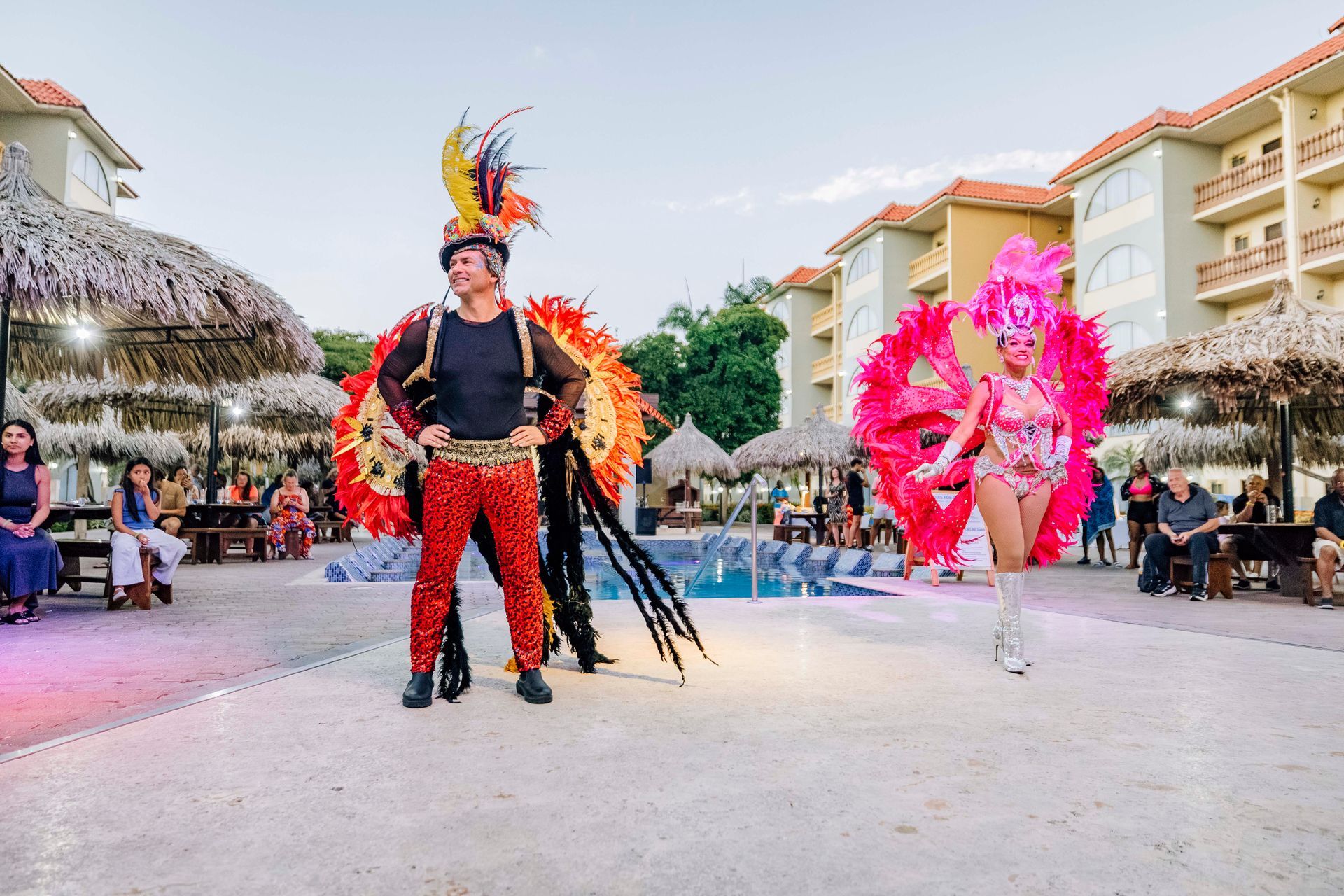 Two performers in colorful costumes pose near a pool with an audience at an outdoor event.