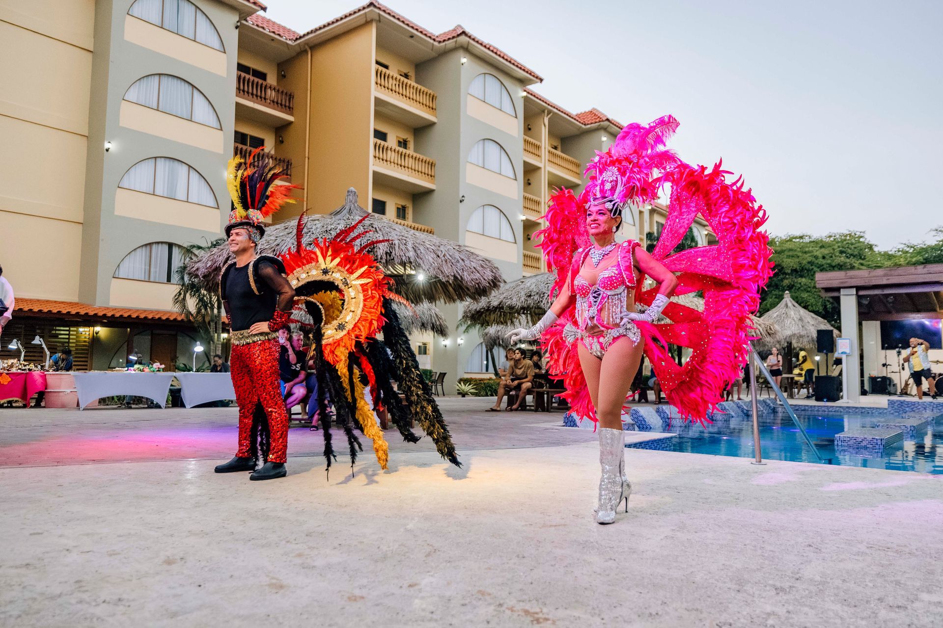 Two people in carnival costumes pose by a pool at an outdoor event.