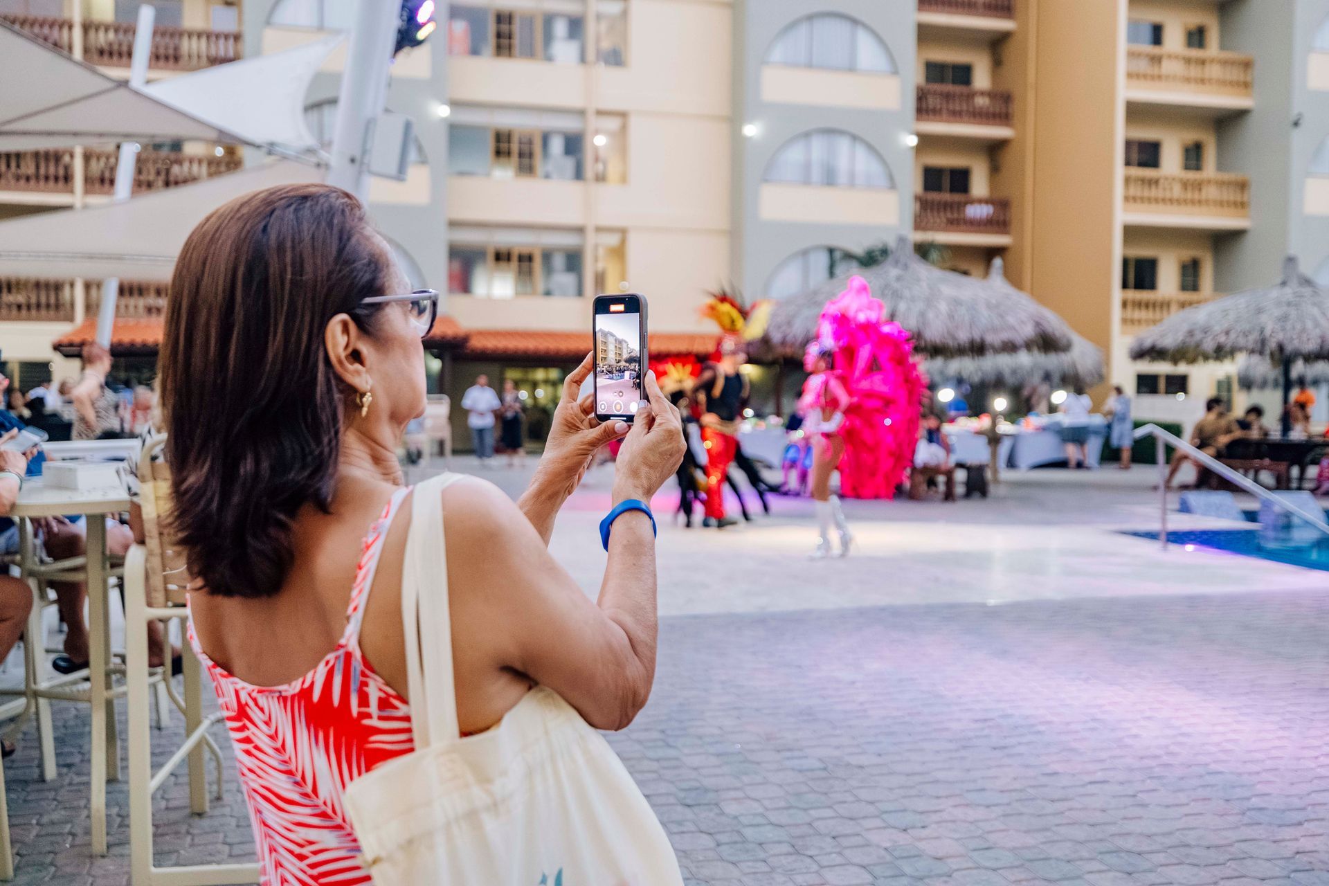 Woman taking a photo with a phone of performers in colorful costumes at an outdoor event near a pool.