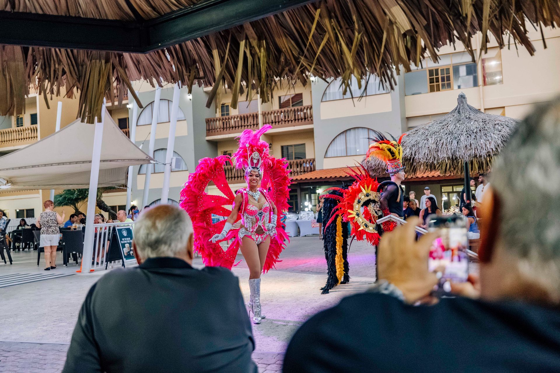 Performers in colorful costumes dancing outdoors. People watch under a thatched roof.