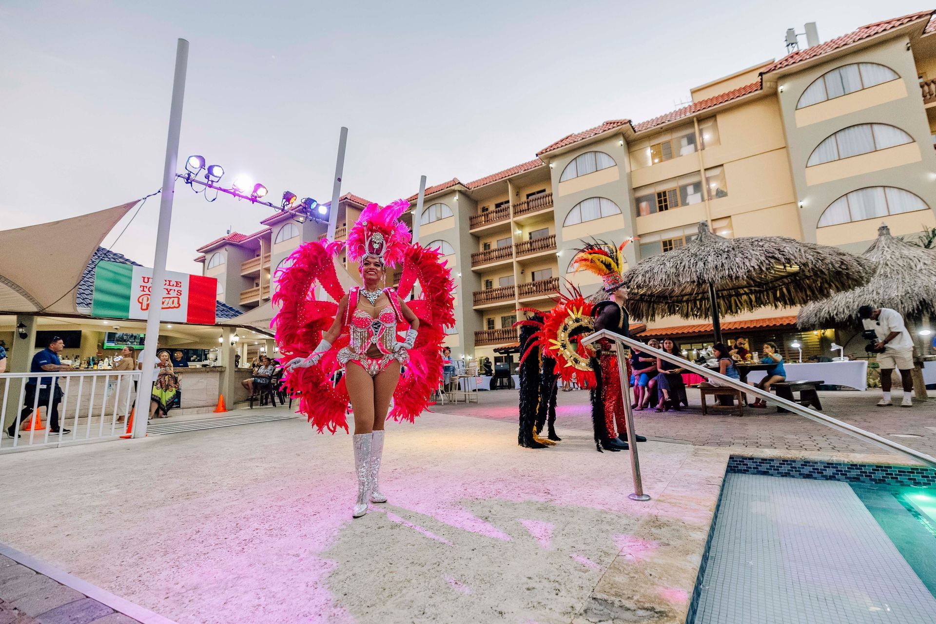 Person in vibrant costume poses by a pool, in front of a multi-story building and event setup.
