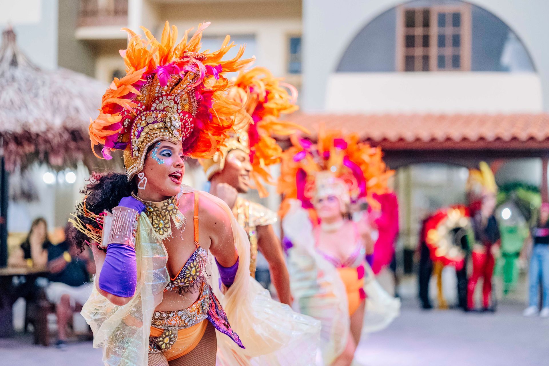 Carnival performers in vibrant costumes, including feathered headdresses, dance outside.