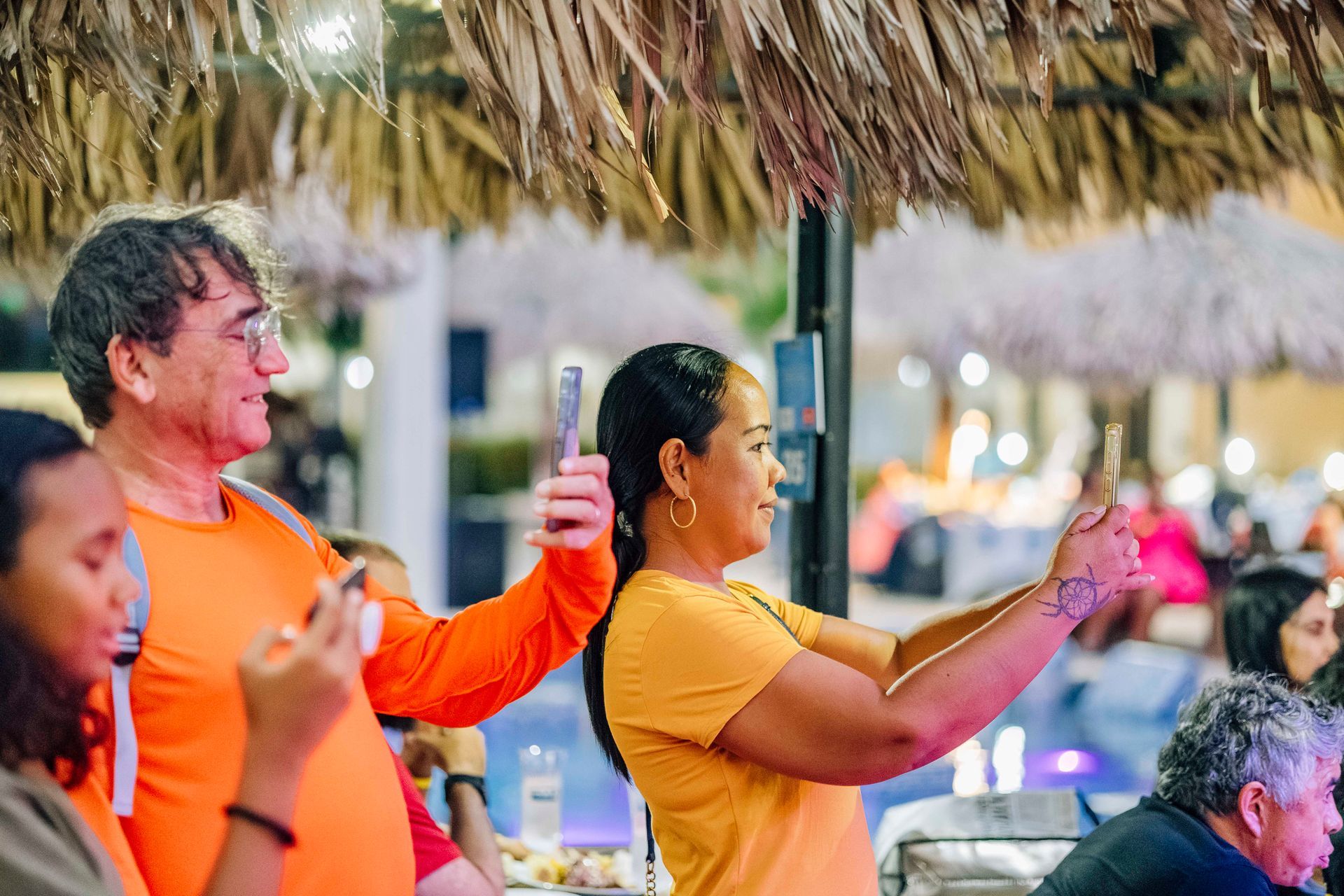 People taking photos with phones near a pool under a thatched roof at night.