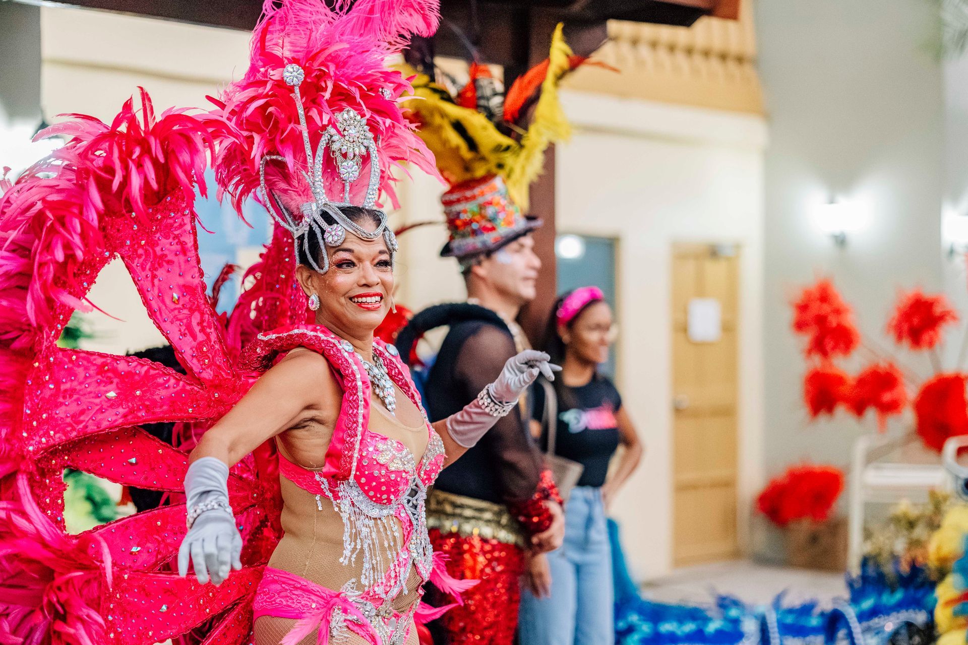 Woman in pink feathered carnival costume smiles and gestures, others in background, indoor event.