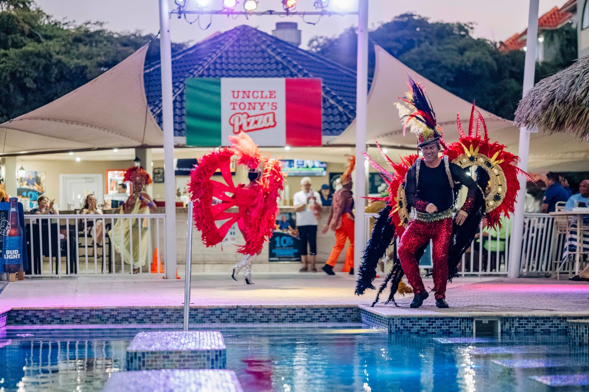 Carnival performers at Uncle Tony's Pizza by a pool; person in red costume dancing.