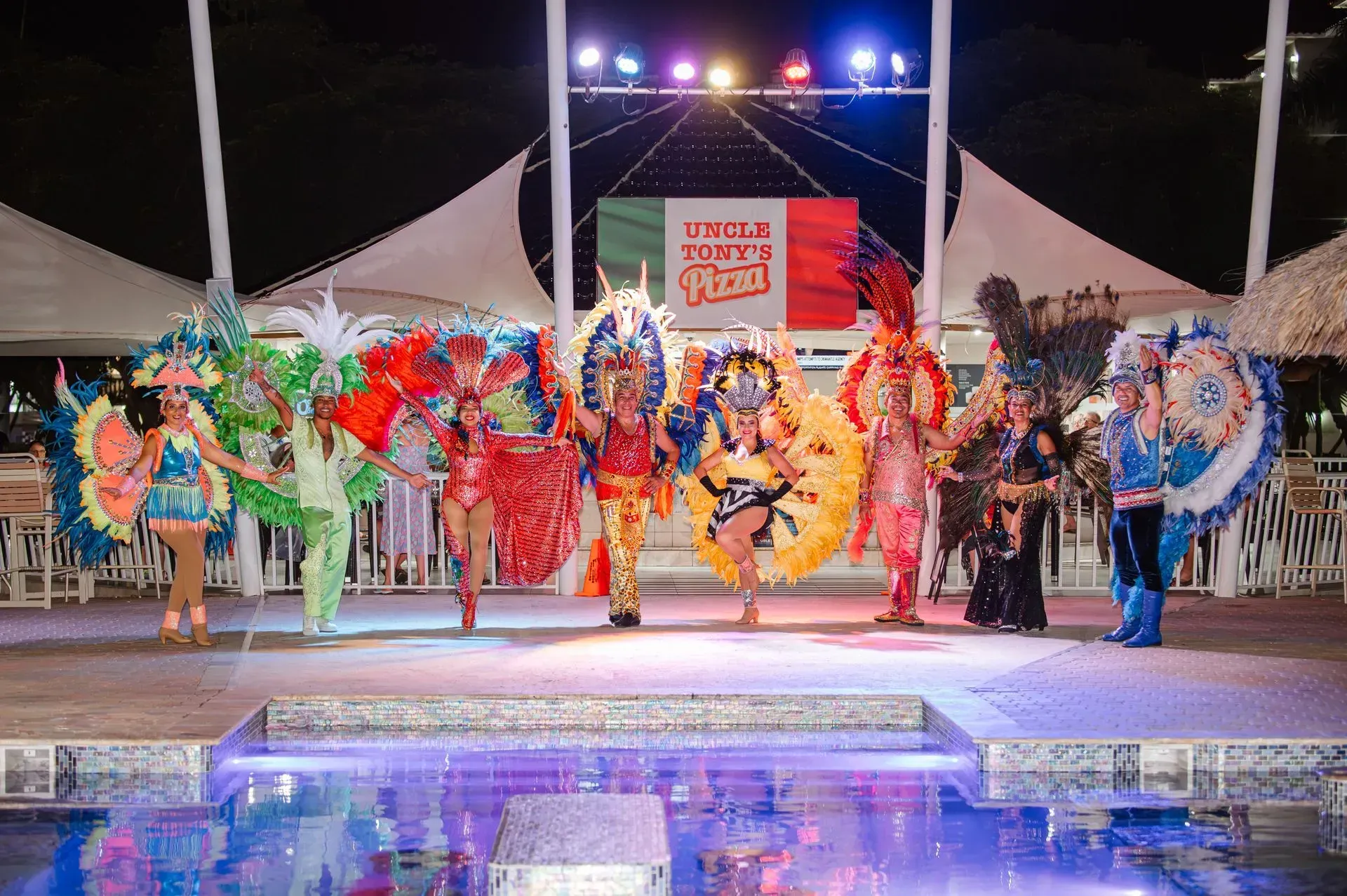 A group of people in colorful costumes are standing on a stage in front of a pool.