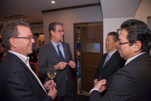 Four men in suits conversing in an indoor setting; one holds a glass of wine, expressions vary.