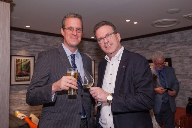 Two men in suits toasting with drinks. One holds a beer, the other wine, in a warmly lit room.