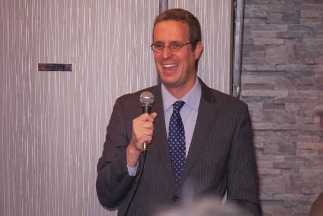 Man in suit smiling, holding a microphone. Speaking in a room with textured walls.