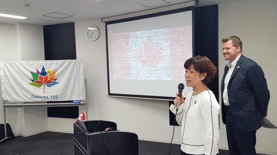 Woman speaking at a podium with a microphone, man standing behind her. Canadian flags are displayed in a room.