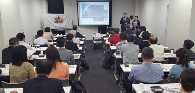A conference with presenters on stage and attendees in rows of seats. A projection screen displays a presentation.