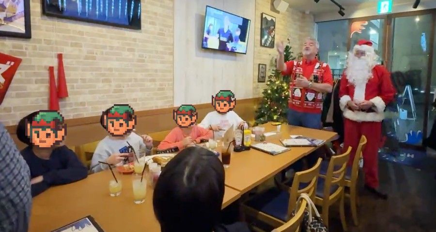 A group of people at a restaurant celebrating Christmas; Santa Claus stands near a man speaking to the group.