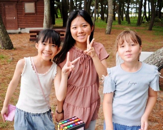 Three girls smiling outdoors, one holding up a peace sign, near trees and a building.