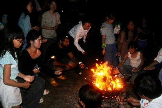 People around a bonfire at night, roasting something on sticks, flames glowing brightly.