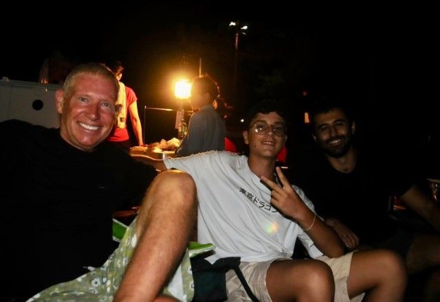 Three men smile at camera in dim outdoor setting; one flashes peace sign.