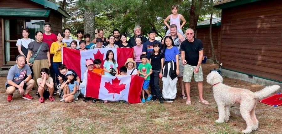 Group of people holding a Canadian flag outside cabins, with a large dog looking on.