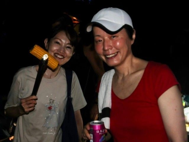 Two smiling women, one holding a corn-on-the-cob with tongs, the other wearing a white cap and holding a can. Dark background.