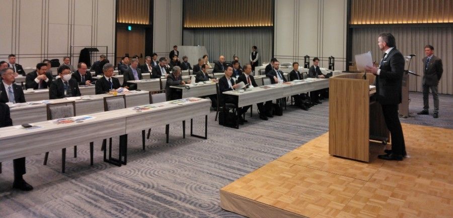 A man speaks at a podium in a conference hall. Attendees sit at tables, listening.