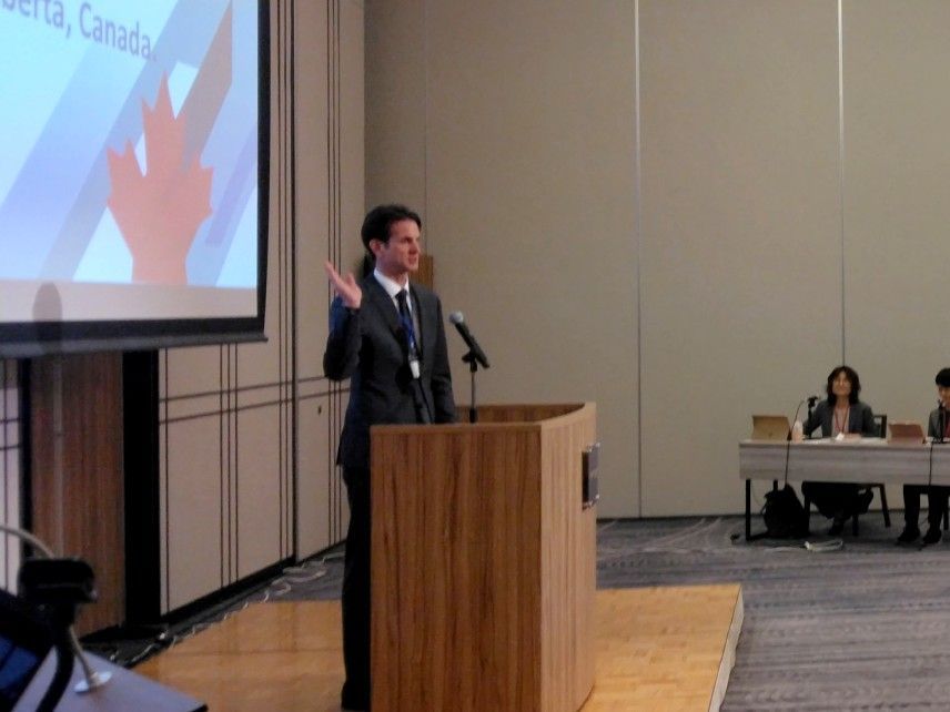Canada Consul in Nagoya speaking at podium, gesturing. Presentation screen behind him; other people sit at a table.