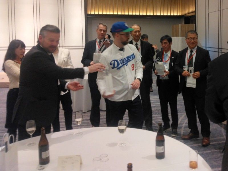 Man in Dodgers jersey and hat participates in a wine tasting. Other people in suits watch.