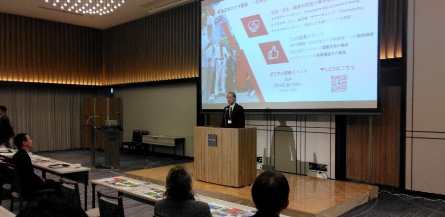 A man giving a presentation at a conference. He is standing at a podium in front of a screen with Japanese text.