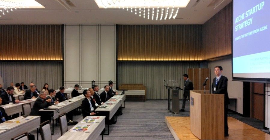 Man giving a presentation at a conference. Attendees seated at tables in a room with a projector screen.
