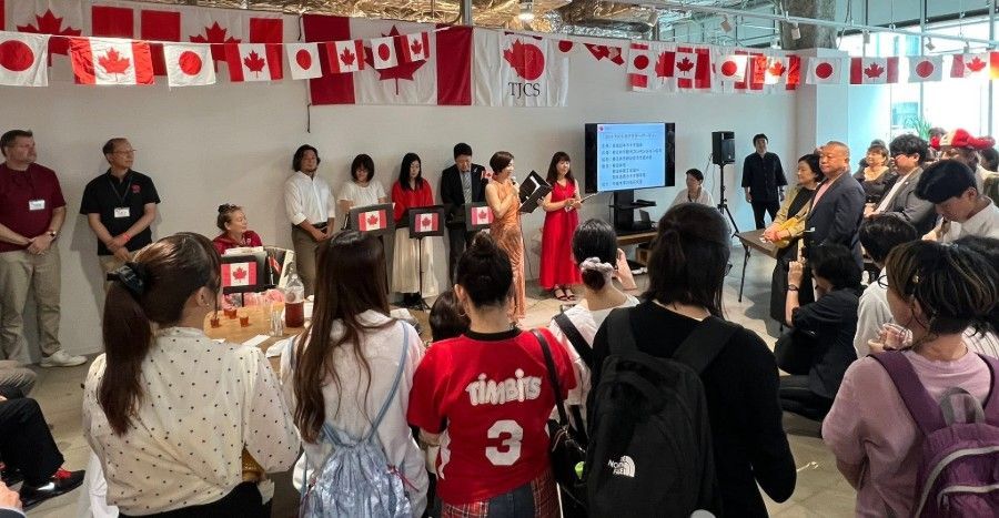 People standing in a circle at a Canada-themed gathering with flags, speakers, and a presentation screen