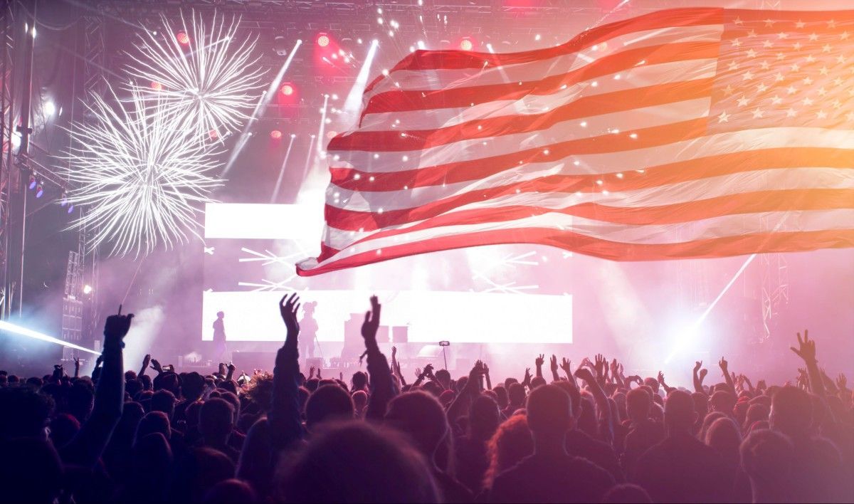 Crowd at a concert with fireworks and a large American flag waving overhead