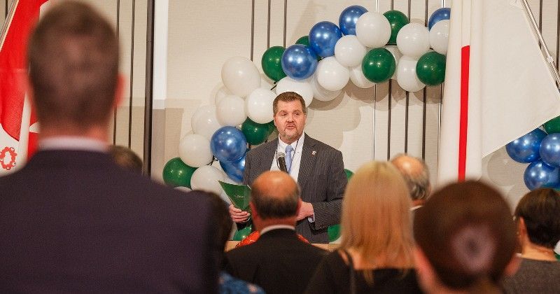Man speaking at an event, standing in front of balloons and flags; audience present.
