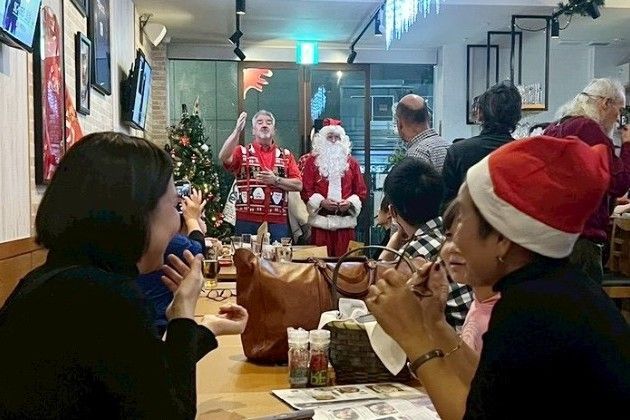 People at a restaurant celebrate Christmas. Santa and an elf-like person stand in the background, festive decorations around.