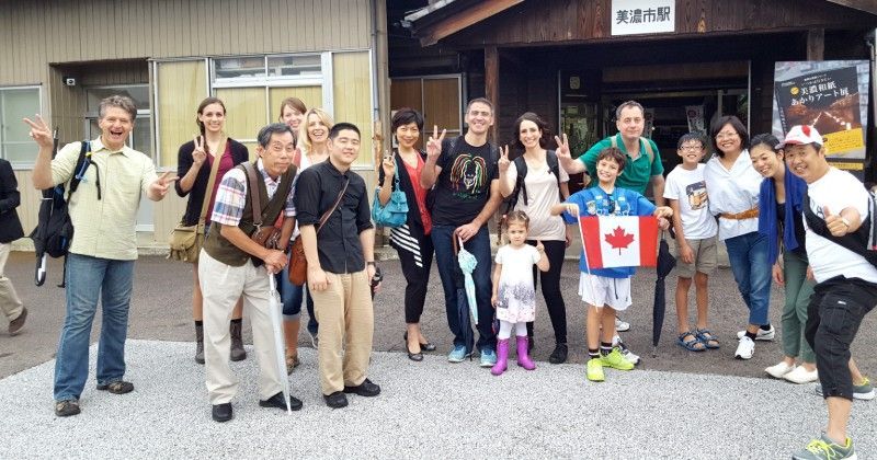 Group of people posing in front of a building; some hold up peace signs. One child has a Canadian flag.