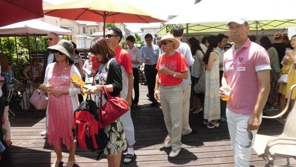 People gathered outdoors under umbrellas. Some wearing hats, dressed casually, socializing at an event.